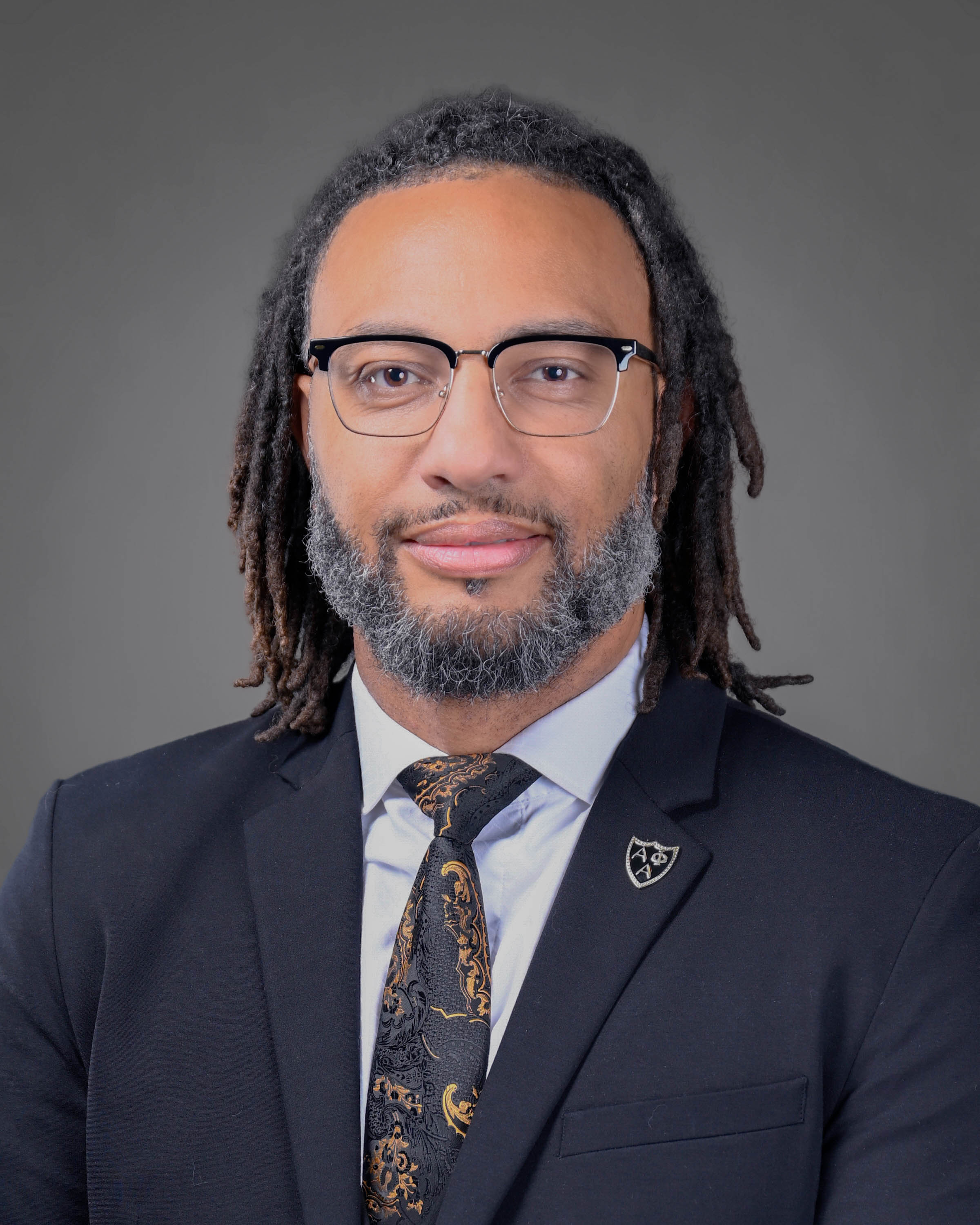 Man with long curly dark hair and a beard in a dark suit with gray shirt and dark tie.